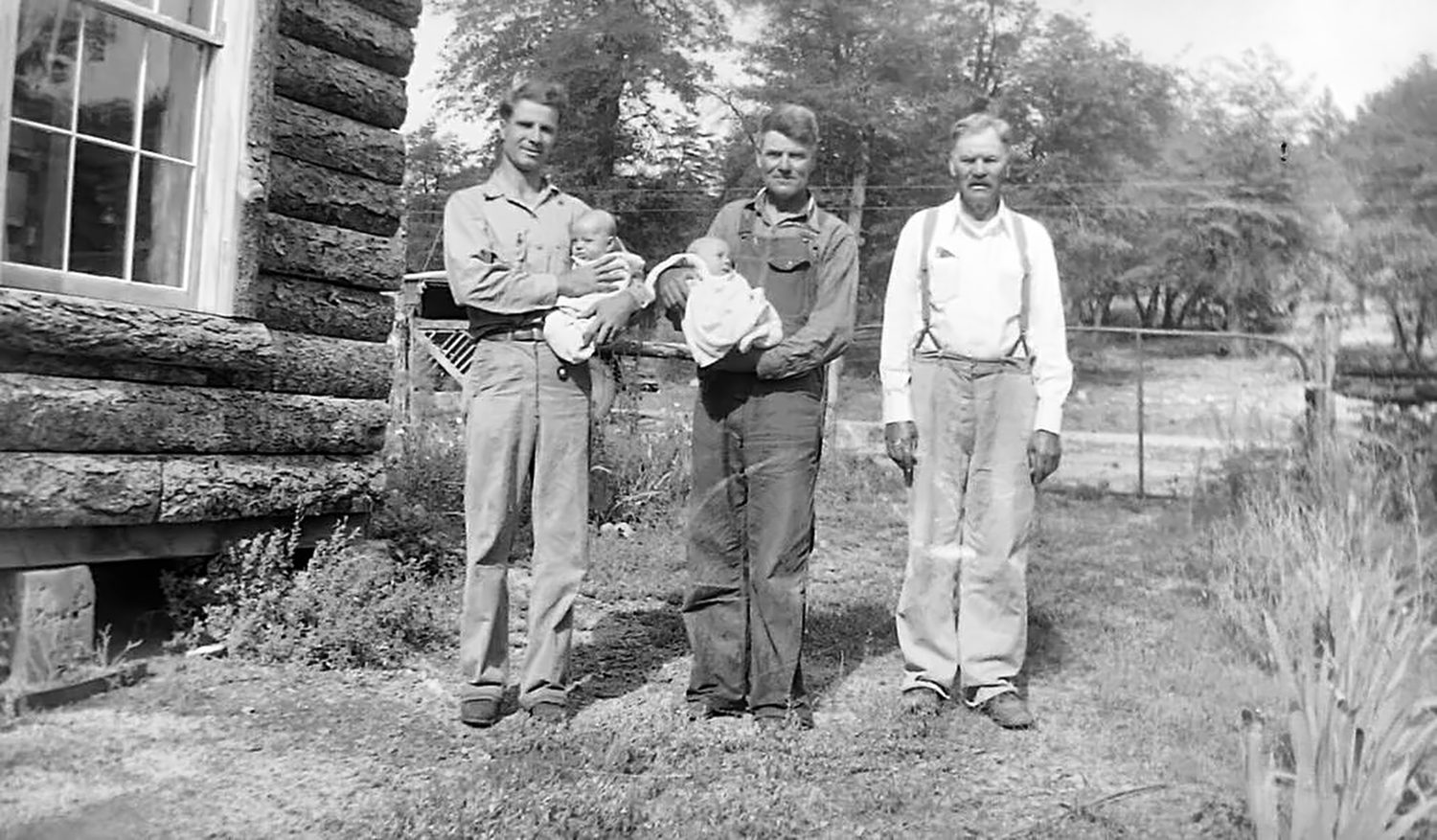 The same session outside a log house: the young man and the man in overalls each hold one twin, and the old man stands at the right in a white shirt and suspenders.