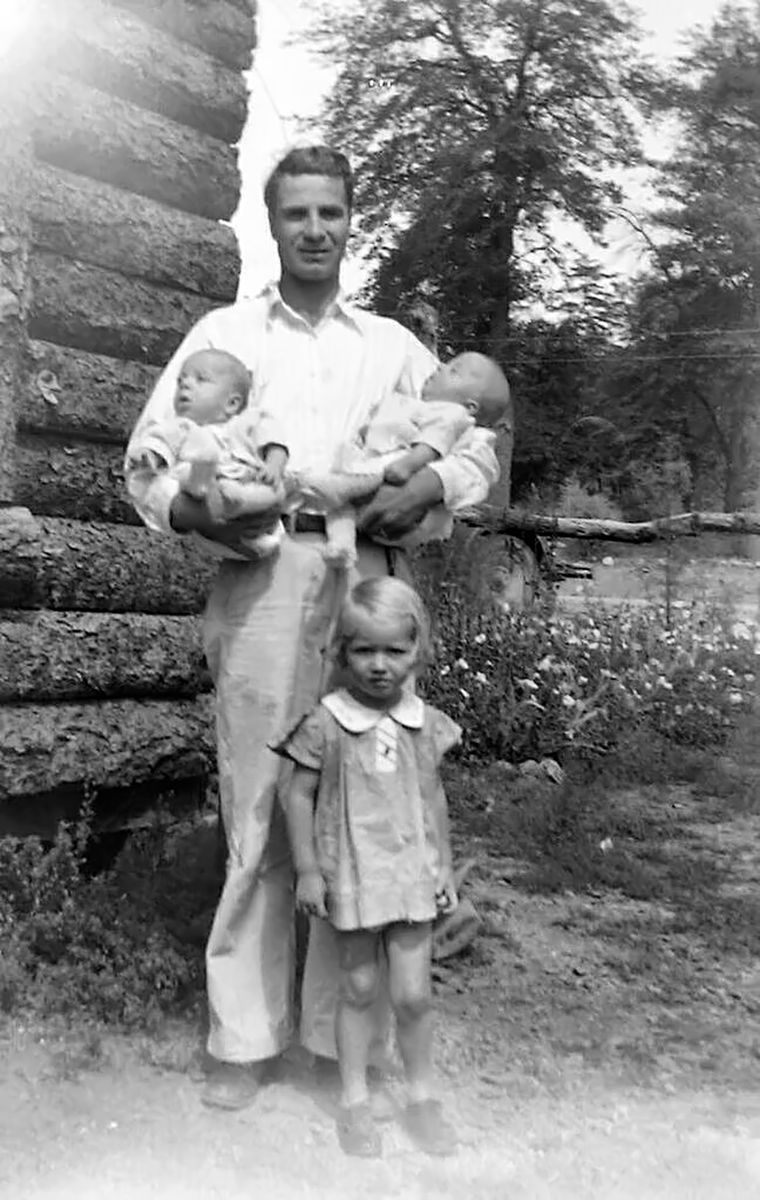 A young man in a white shirt holds two infants in front of a log wall; a small fair-haired girl in a dress stands in front of him.