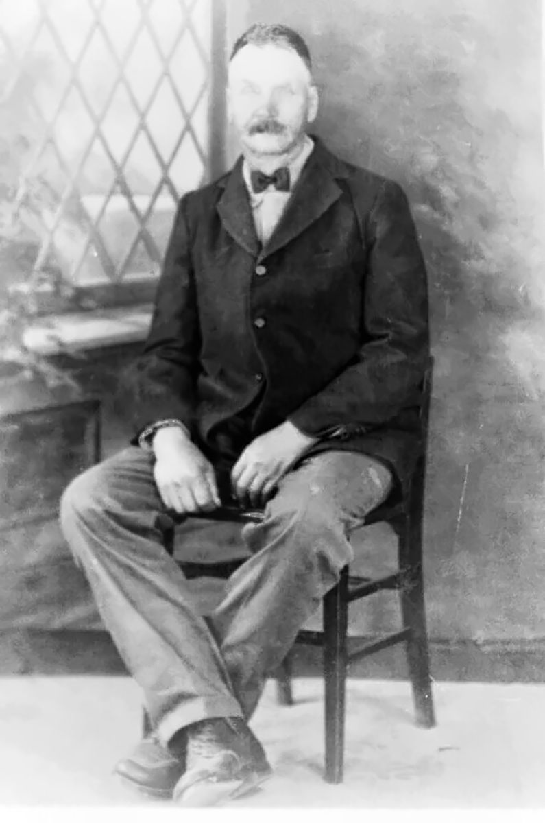A seated studio portrait of a man with a heavy moustache in a dark sack coat, bow tie and light trousers, hands on his knees, a lattice window painted on the backdrop behind him.