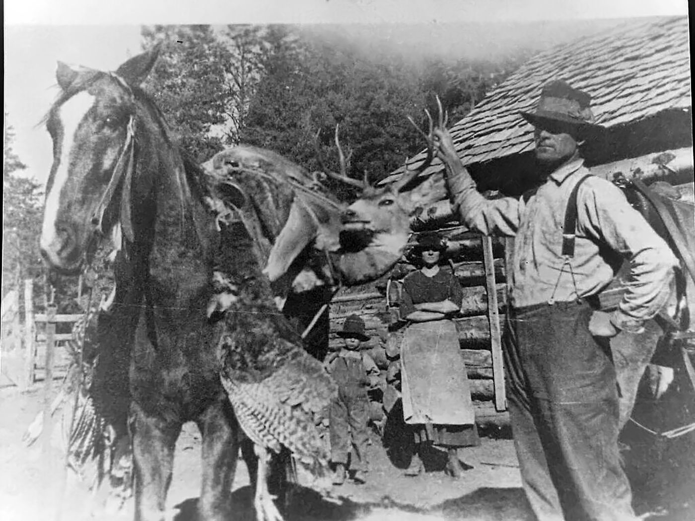 A man in a hat, suspenders and work shirt stands beside a horse packed with a buck deer and a wild turkey, in front of a log house; a woman in an apron and a small boy stand in the doorway.